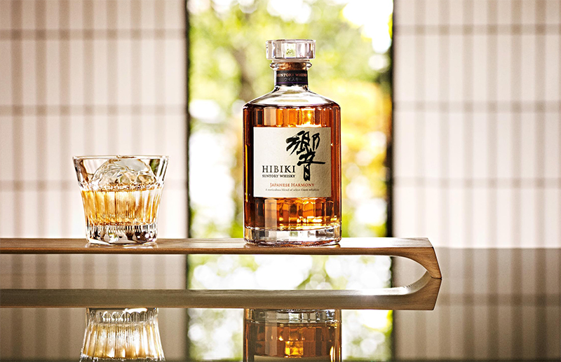 Hibiki on the Rocks im Glas mit einem Eiswürfel, neben einer vollen Flasche Hibiki auf einem Holzbrett platziert auf einem Tisch