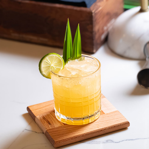 Laphroaig Smoked Pineapple Mule Cocktail mit 2 Ananas-Blättern und einer Limetten-Scheibe garniert, auf einem Holzbrett, platziert auf einem Marmor-Tisch