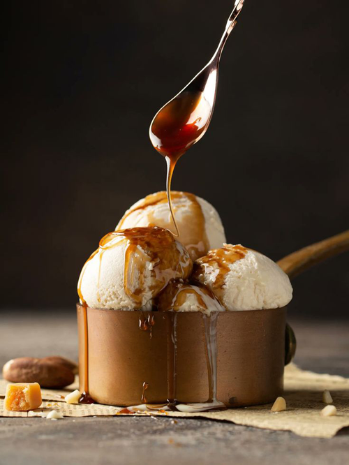 3 Kugeln Vanilleeis mit karamellisierten Pekannüssen und Karamelsoße, die mit einem Löffel darauf geträufelt wird in einem Metallbecher