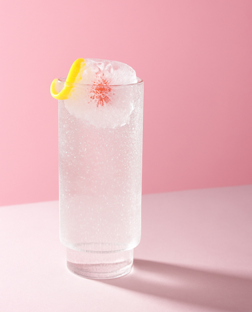 ROKU Summer Bloom Cocktail mit einem infused Ice Cube mit Kirschblütenblättern und Zitronenzeste vor einem rosa-farbenen Hintergrund