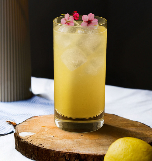 Roku Yuzu Spark Cocktail mit Essbaren Blütetn im Glas auf einer Holzscheibe neben einer Zitrone