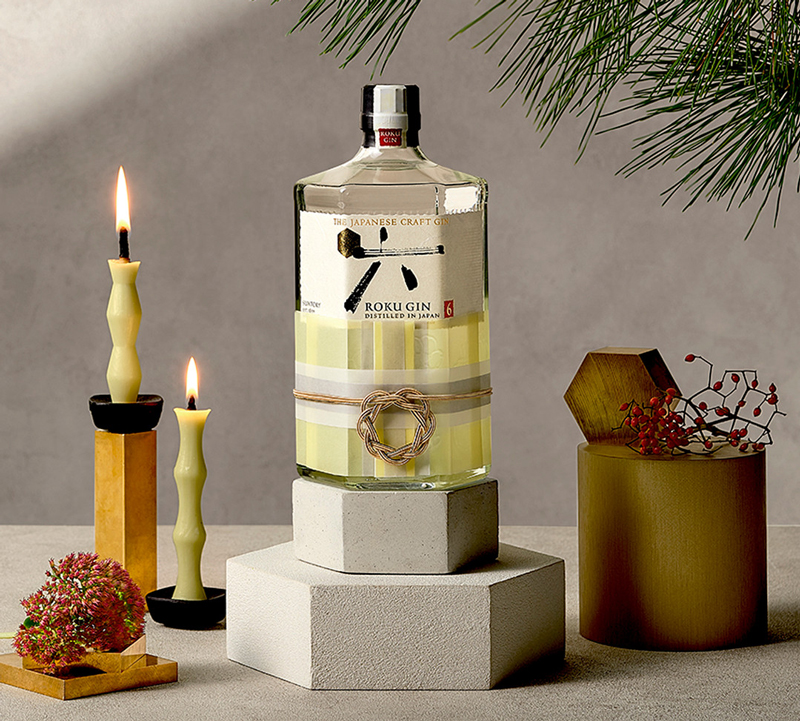 Eine Flasche Roku Gin auf einem Beton-Podest neben zwei angezündeten Kerzen und Deko unter einem Tannenzweig