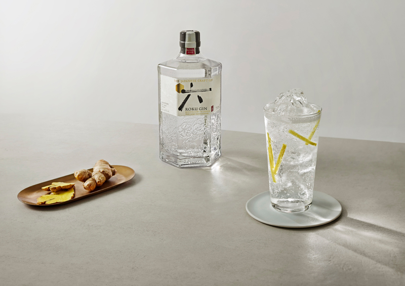 Ein Glas Gin & Tonic mit Ingwerstäbchen neben einer Flasche Roku Gin und einem Teller mit aufgeschnittenem Ingwer und einer Ingwerwurzel
