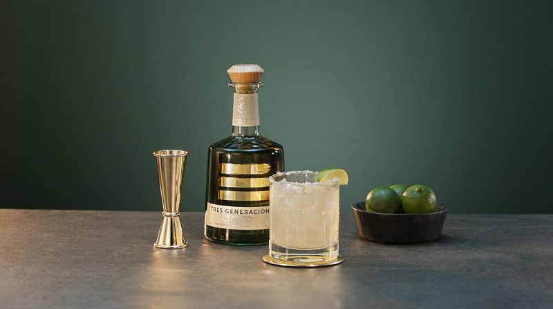 Tres Margarita Cocktail garniert mit Zuckerrand und Limettenviertel neben einer Flasche Tres Generaciones Reposado, einem Messebecher und einer Schale mit 3 Limetten