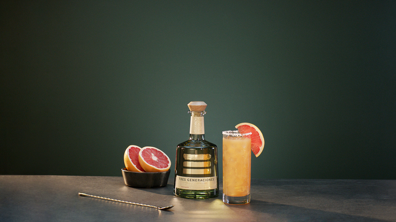 Tres Paloma Cocktail im Glas garniert mit einer Grapefruit-Scheibe neben einer Flasche Tres Generaciones Reposado, einer Schale mit aufgeschnittenen Grapefruits und einem Löffel