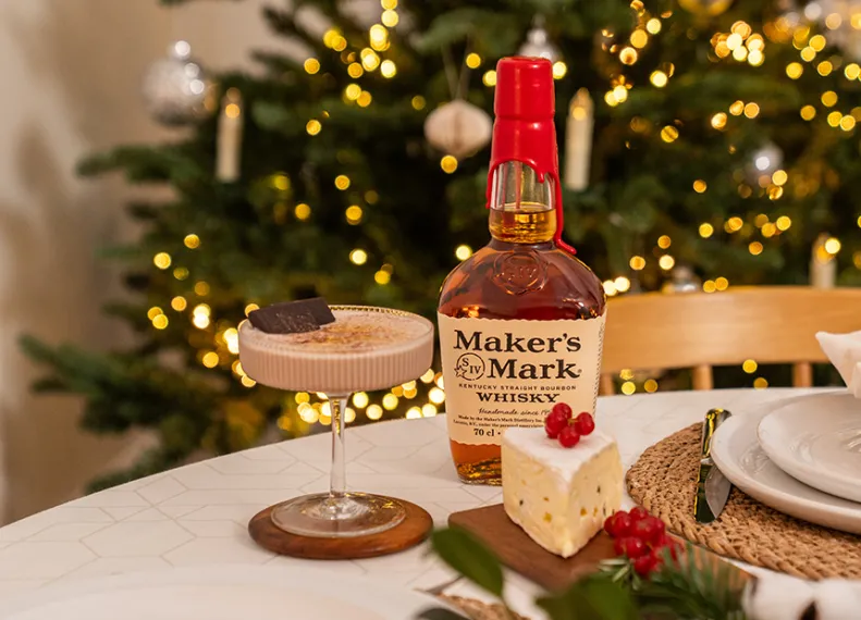 Maker's Mark Bourbon und Cocktail auf einem festlich gedeckten Tisch mit Teller vor einem geschmückten Weihnachtsbaum