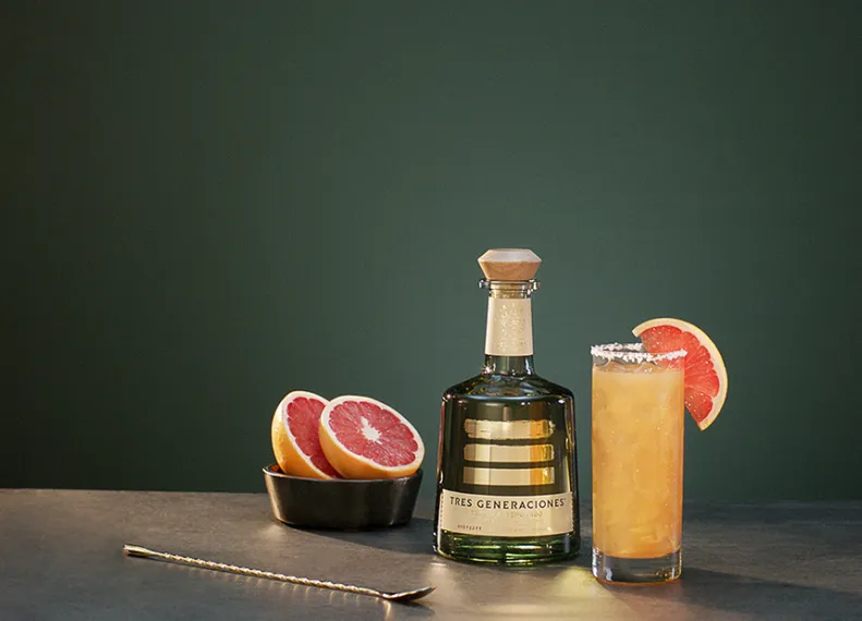 Tres Paloma Cocktail im Glas garniert mit einer Grapefruit-Scheibe neben einer Flasche Tres Generaciones Reposado, einer Schale mit aufgeschnittenen Grapefruits und einem Löffel