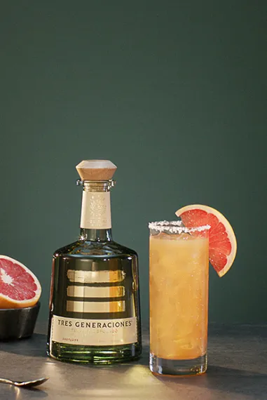 Tres Paloma Cocktail im Glas garniert mit einer Grapefruit-Scheibe neben einer Flasche Tres Generaciones Reposado, einer Schale mit aufgeschnittenen Grapefruits und einem Löffel
