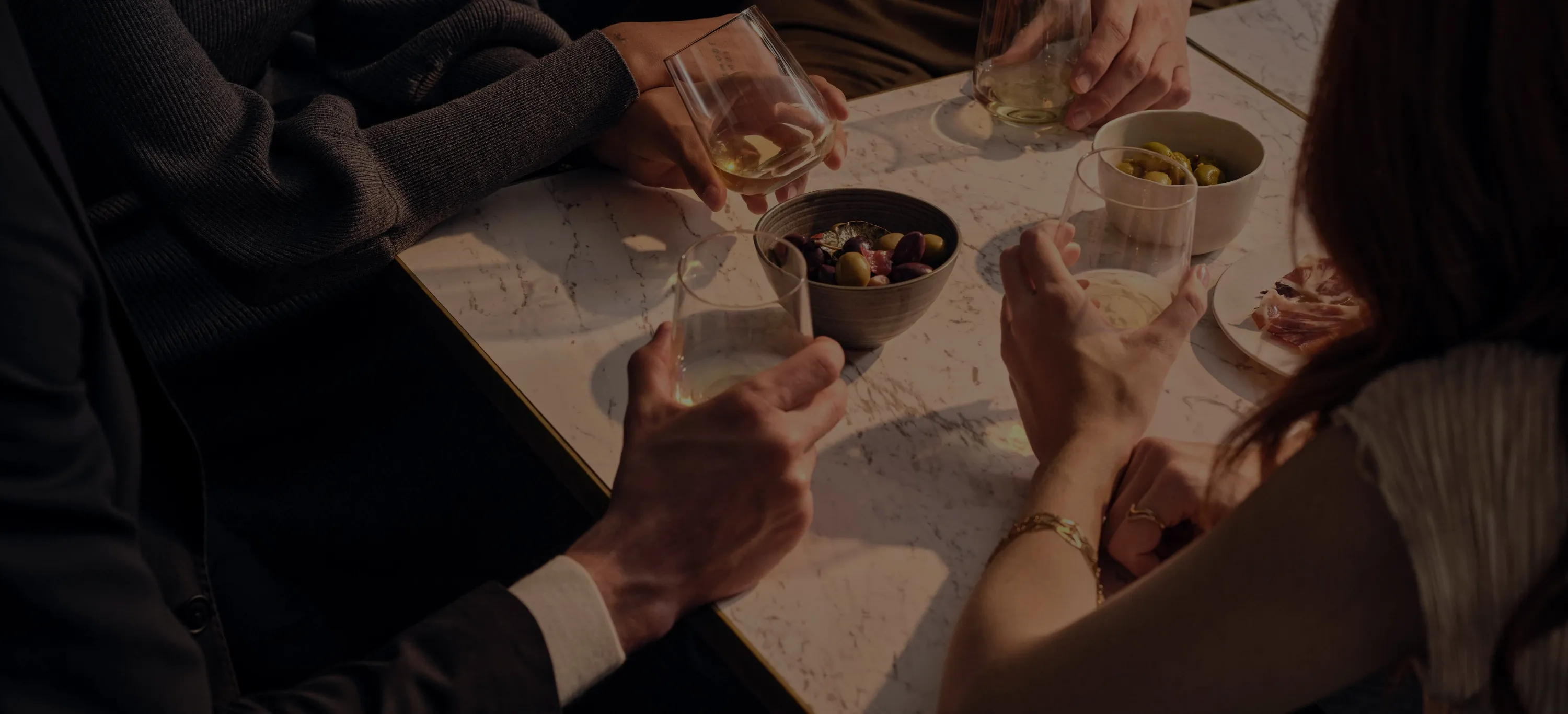 Four people holding glasses, sat around a table. A bowel of olives and a plate of cold meats beside them. 