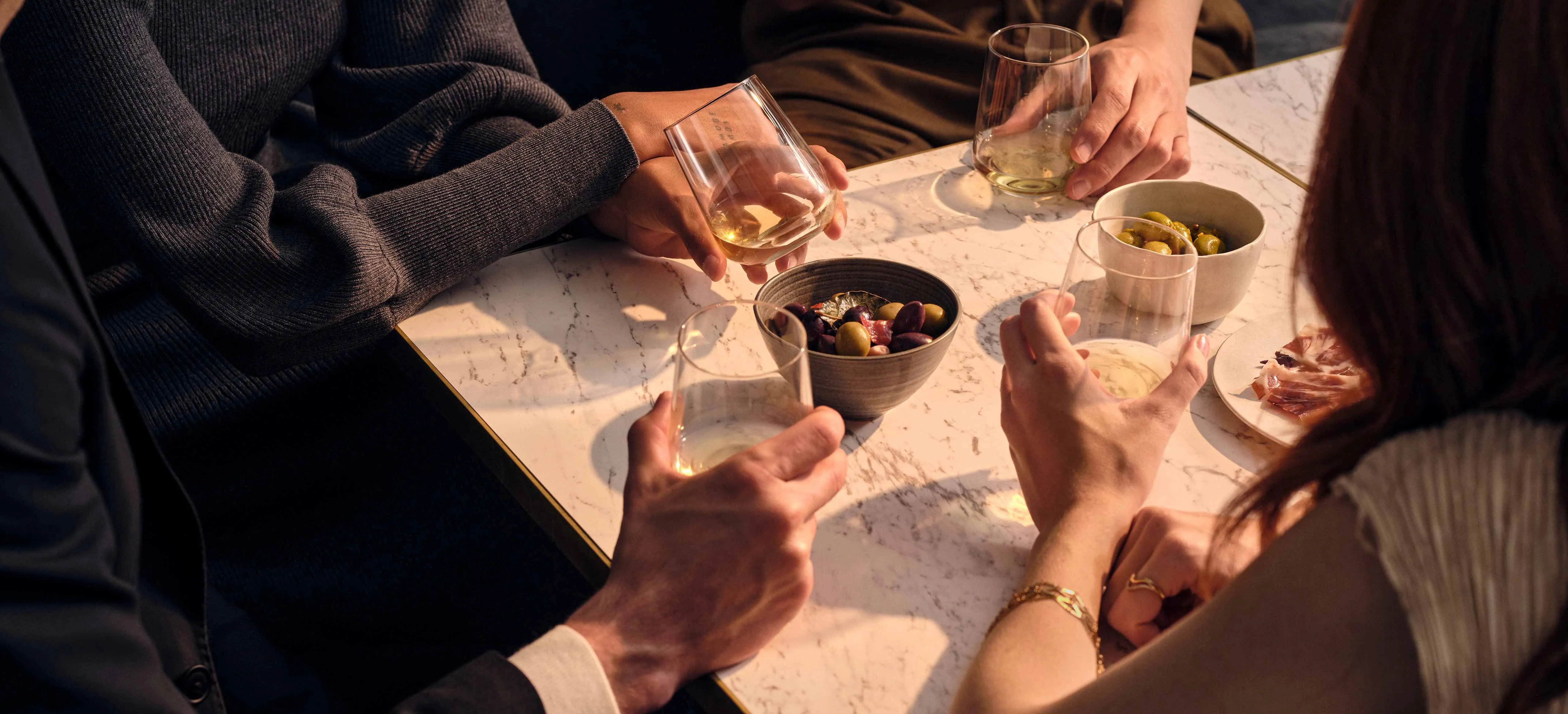 Four people holding glasses, sat around a table. A bowel of olives and a plate of cold meats beside them.
