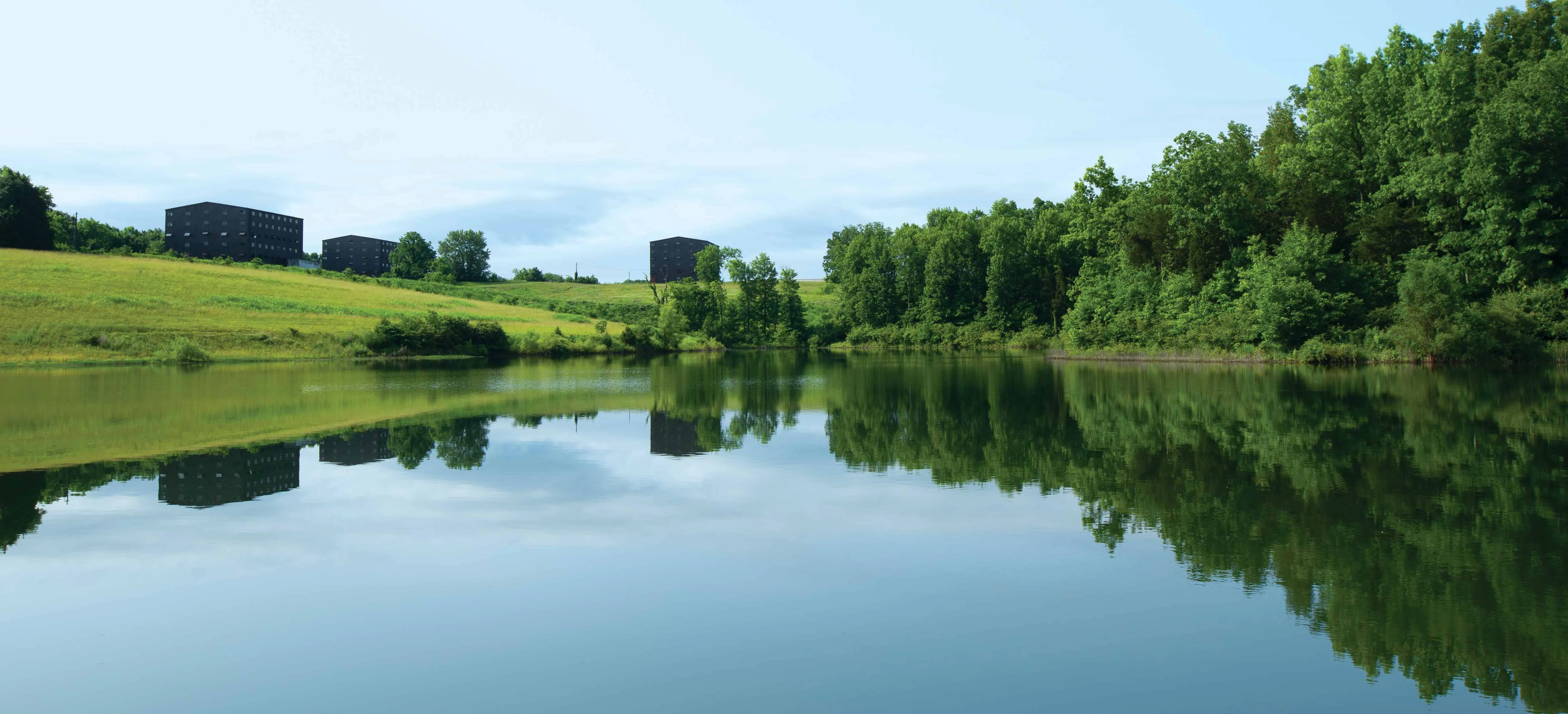 A wide expanse of water showing the reflection of trees and the sky. Beyond the banks of the lake are some large distillery buildings.