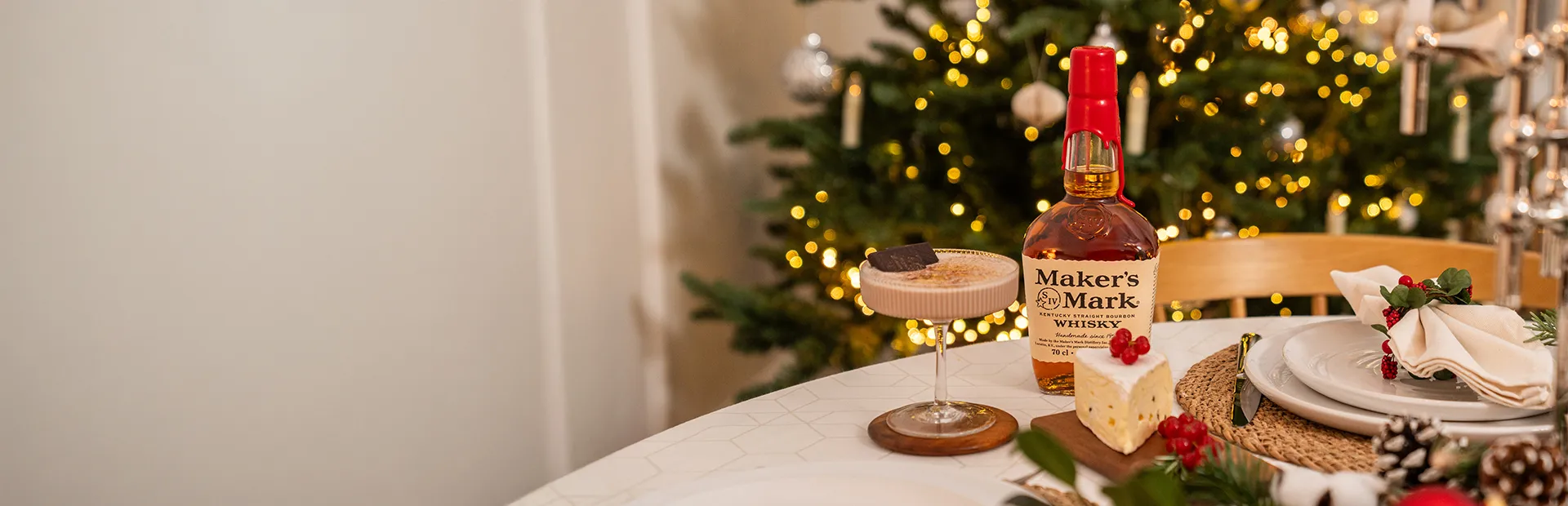 Maker's Mark Bourbon und Cocktail auf einem festlich gedeckten Tisch mit Teller vor einem geschmückten Weihnachtsbaum