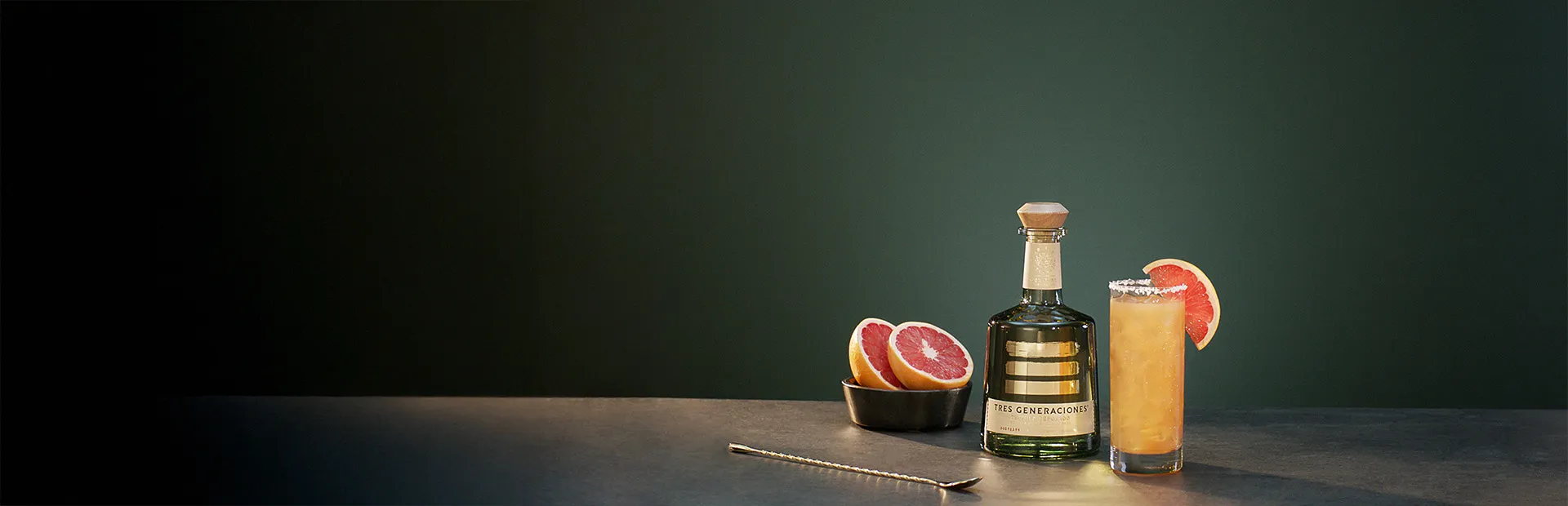 Tres Paloma Cocktail im Glas garniert mit einer Grapefruit-Scheibe neben einer Flasche Tres Generaciones Reposado, einer Schale mit aufgeschnittenen Grapefruits und einem Löffel